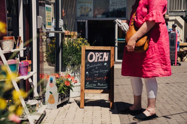 Woman standing in front of one of the shipping container shops, there's an A frame chalk board with 'Open' written on it.