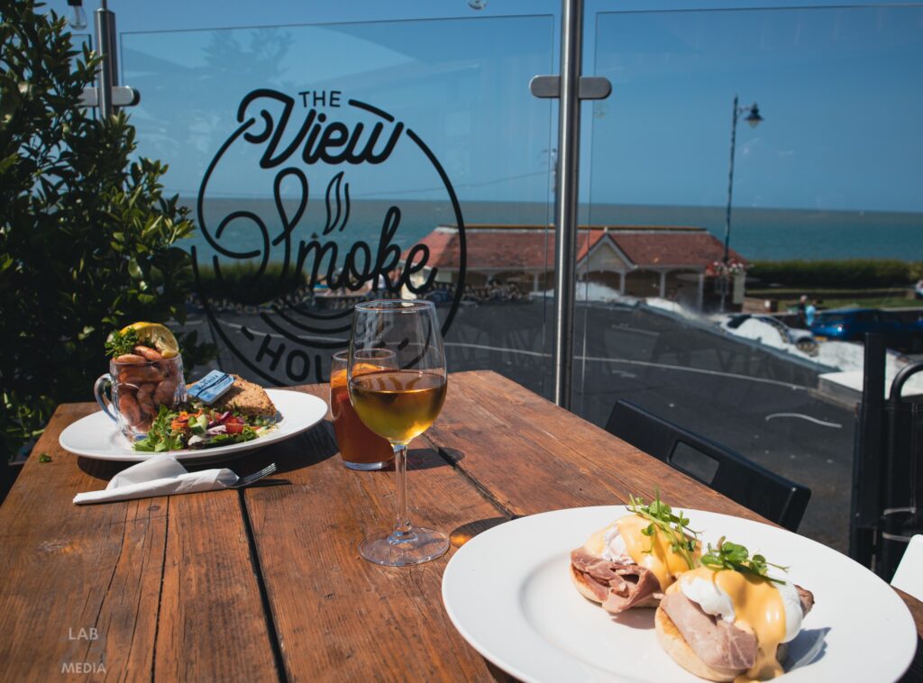 A table with two plates, a glass of white wine, and a glass of beer. Fish dishes on each place. These are placed on a wooden table on the balcony of The View Smokehouse., which overlooks the seafront.