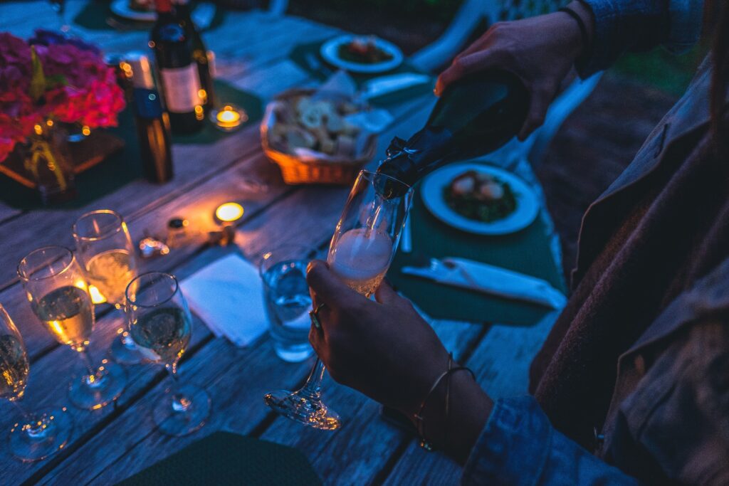 Someone pouring a glass of champagne into a glass whilst sitting at a festive table outdoors at dusk